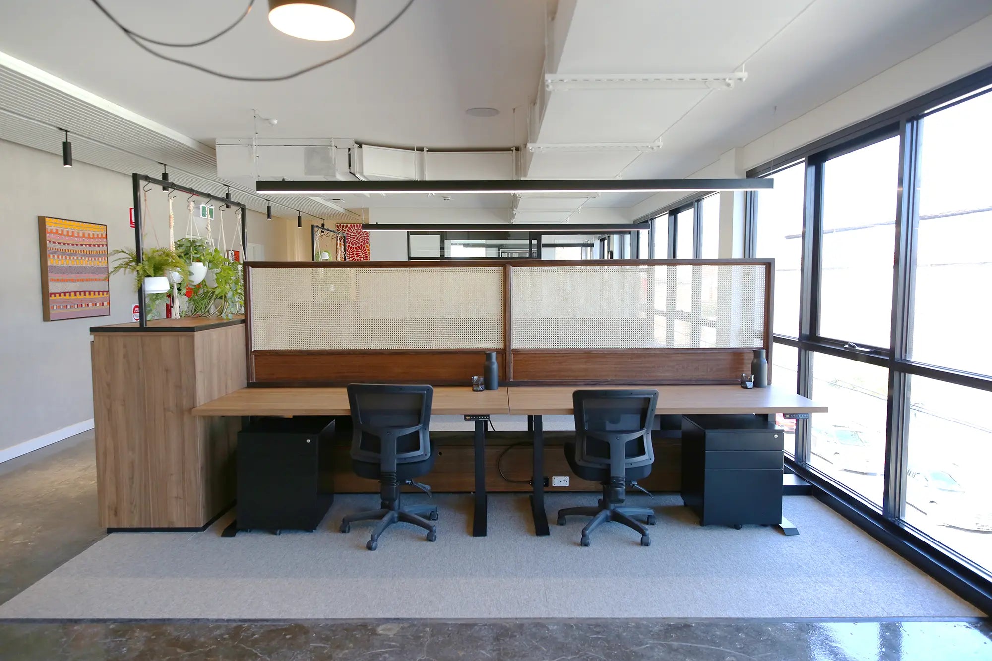 The workspace includes Stando sit-and-stand desks, black ergonomic chairs, and rattan screens, blending functionality with natural design elements.